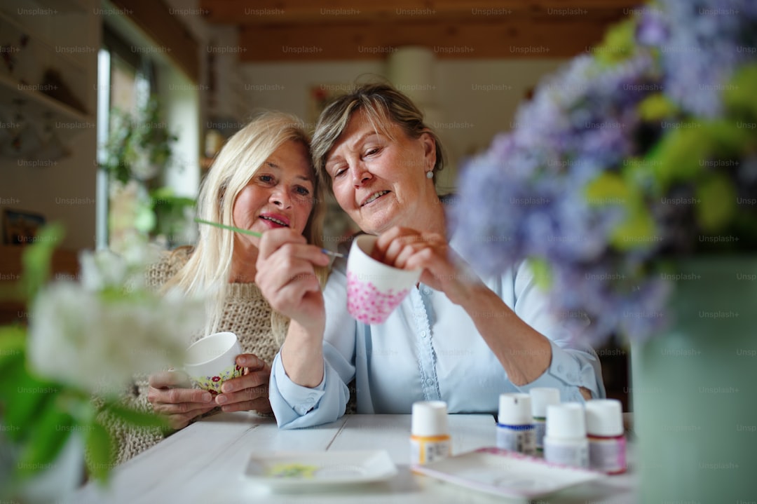 A happy senior woman artist teaching her friend how to paint cermaic cup, artcraft concept.