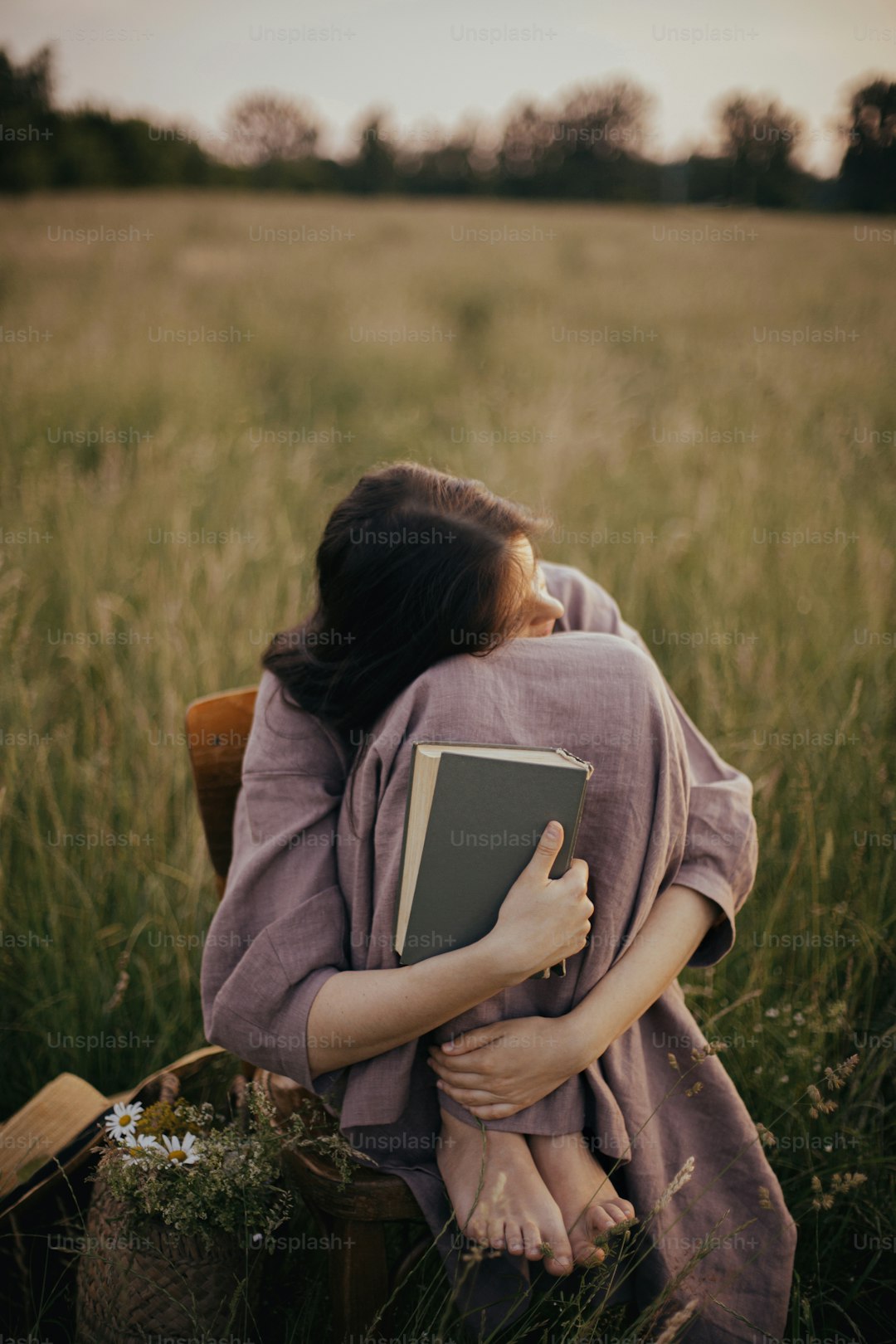 Beautiful woman in linen dress sitting on rustic chair and dreaming in summer meadow. Young female relaxing with book and basket of flowers in countryside sunset. Atmospheric tranquil moment