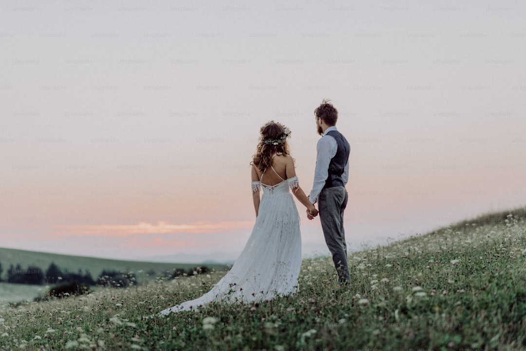 Beautiful young bride and groom outside in green nature at romantic sunset, holding hands. Rear view.