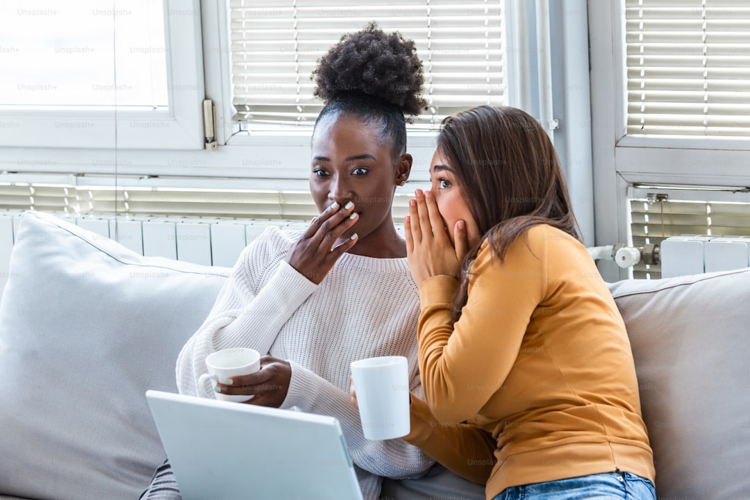 Diverse female friends sharing secrets. Two women gossiping at home. Excited emotional girl whispering to her friend ear while sitting in living-room