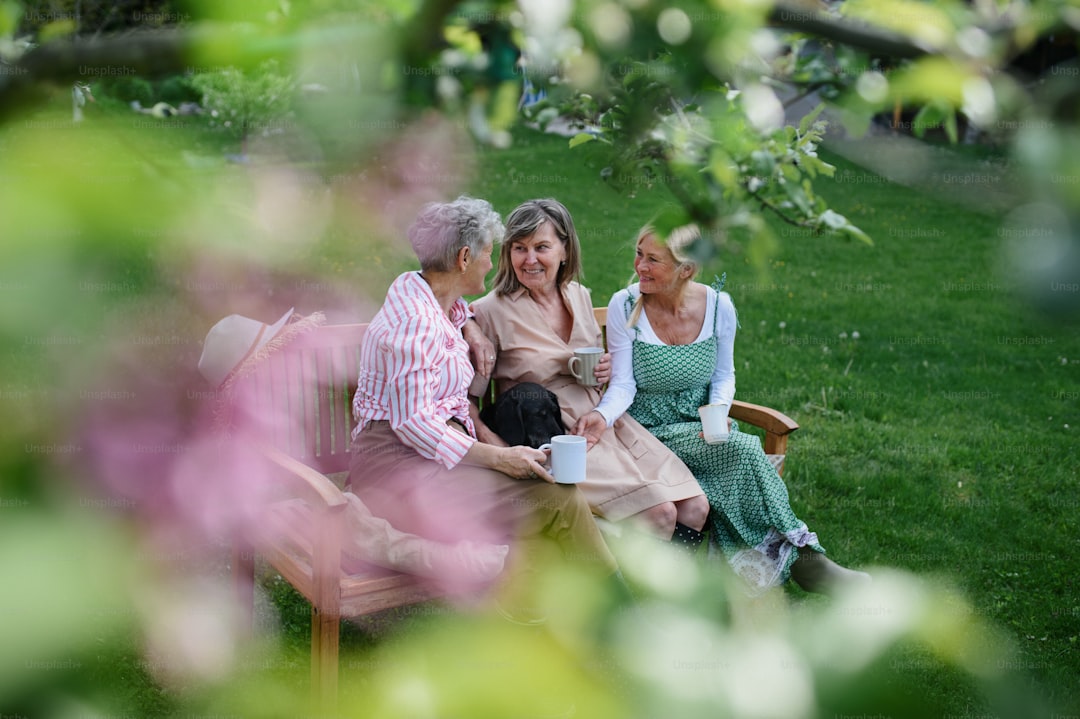 Happy senior women friends sitting on a bench and drinking tea outdoors in garden, laughing.