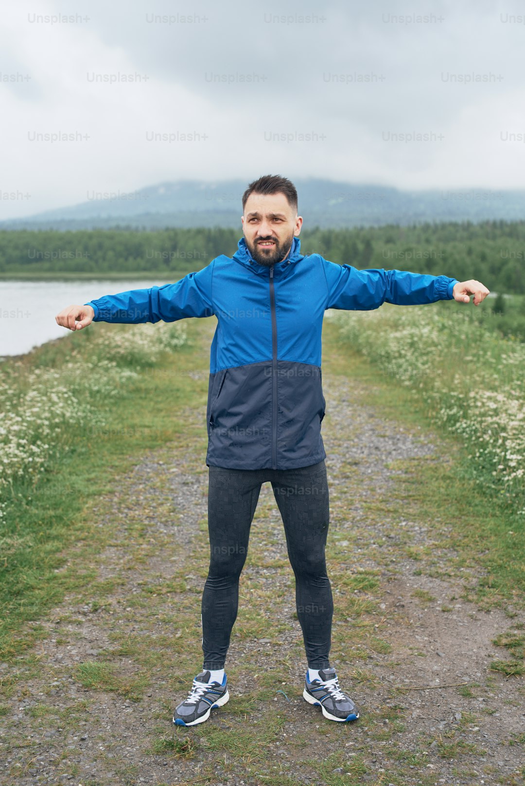 Mid age sportsman exercising outdoor in summer, on gloomy day, standing at the road with scenic view, doing warm up stretching, wearing blue jacket, he has a beard