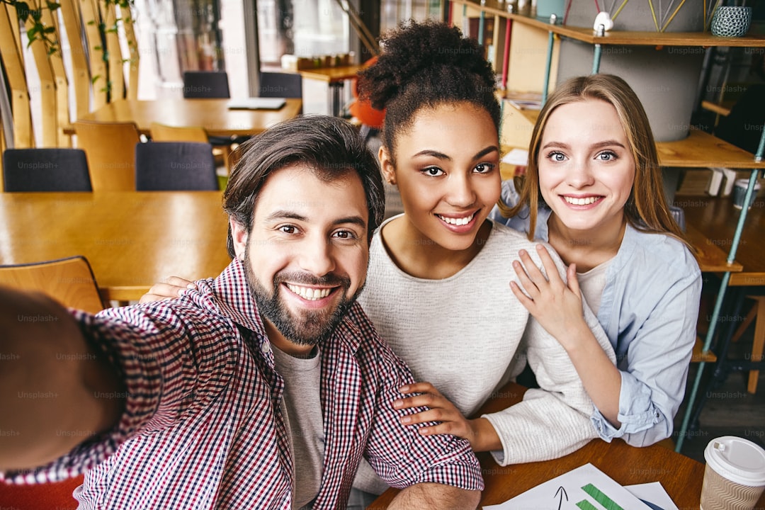 Three laughing young business people in casual wear smile to make shot of themselves. Office interior, bookshelves in the background. Concept of success