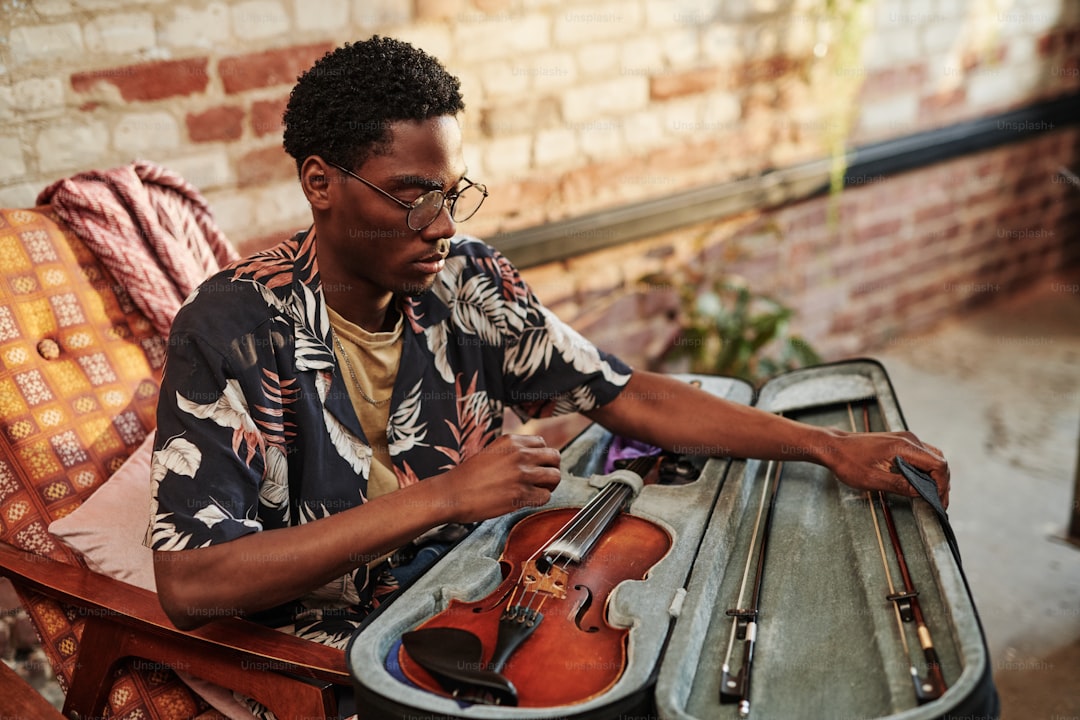 Young black man in stylish casualwear opening slipcover with violin and fiddlesticks while sitting in armchair in living room of loft apartment