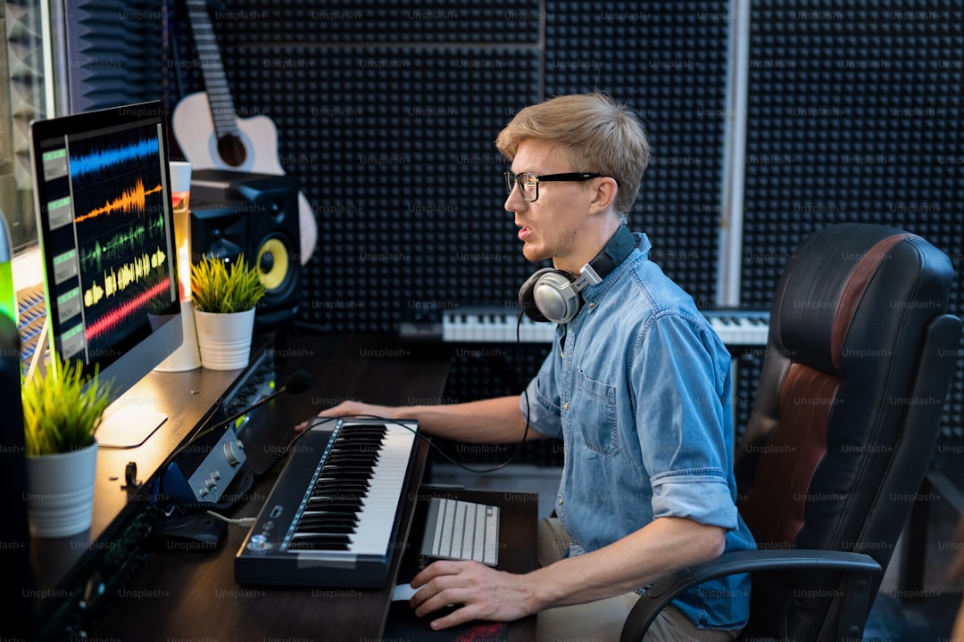 Young male deejay in denim shirt looking at sound mixing tracls on computer screen while sitting by desk in front of monitor